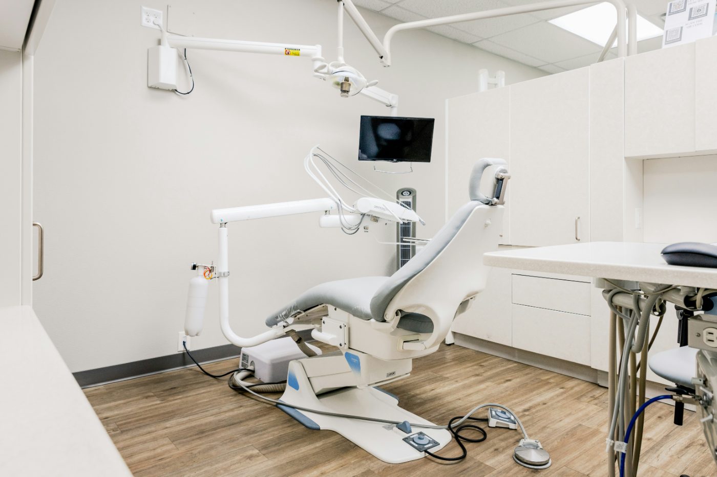 An empty dental treatment room with a gray patient chair, an overhead light and a wall-mounted monitor.
