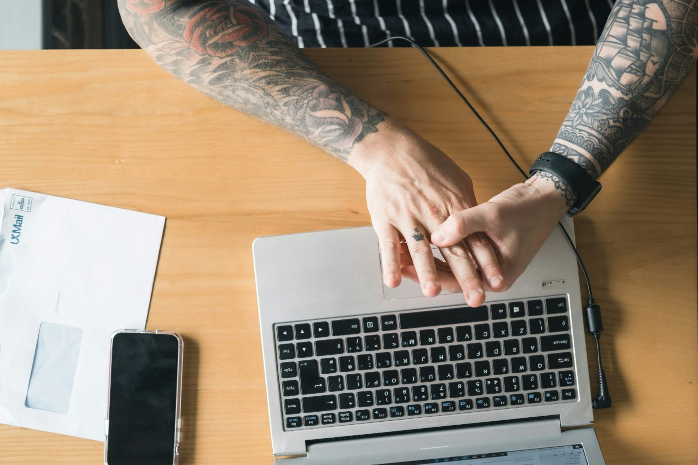 Tattooed forearms and hands resting on a laptop on a wooden table, beside a phone and an envelope.